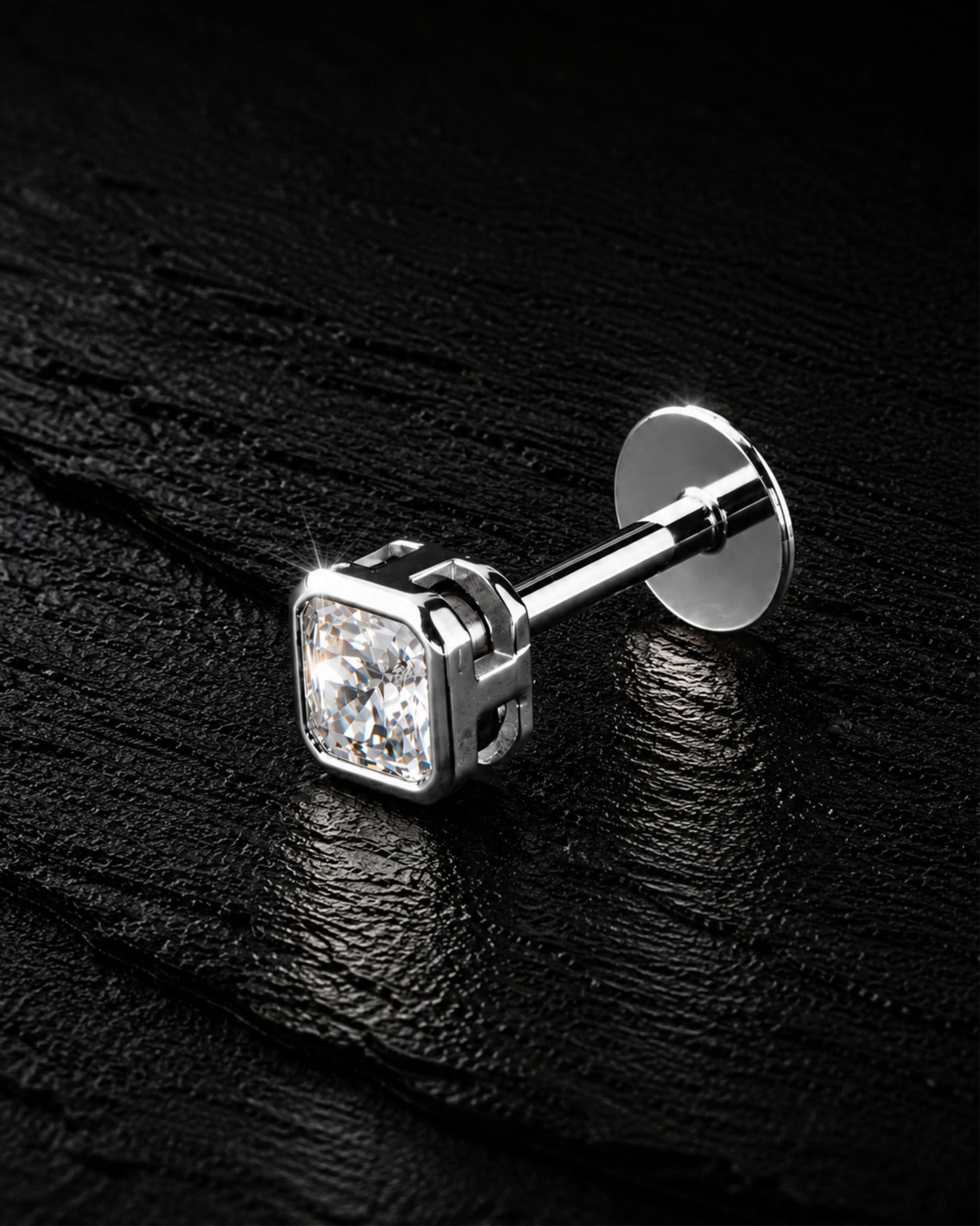 A single silver stud earring with a square-cut clear gemstone, displayed on a textured black background.