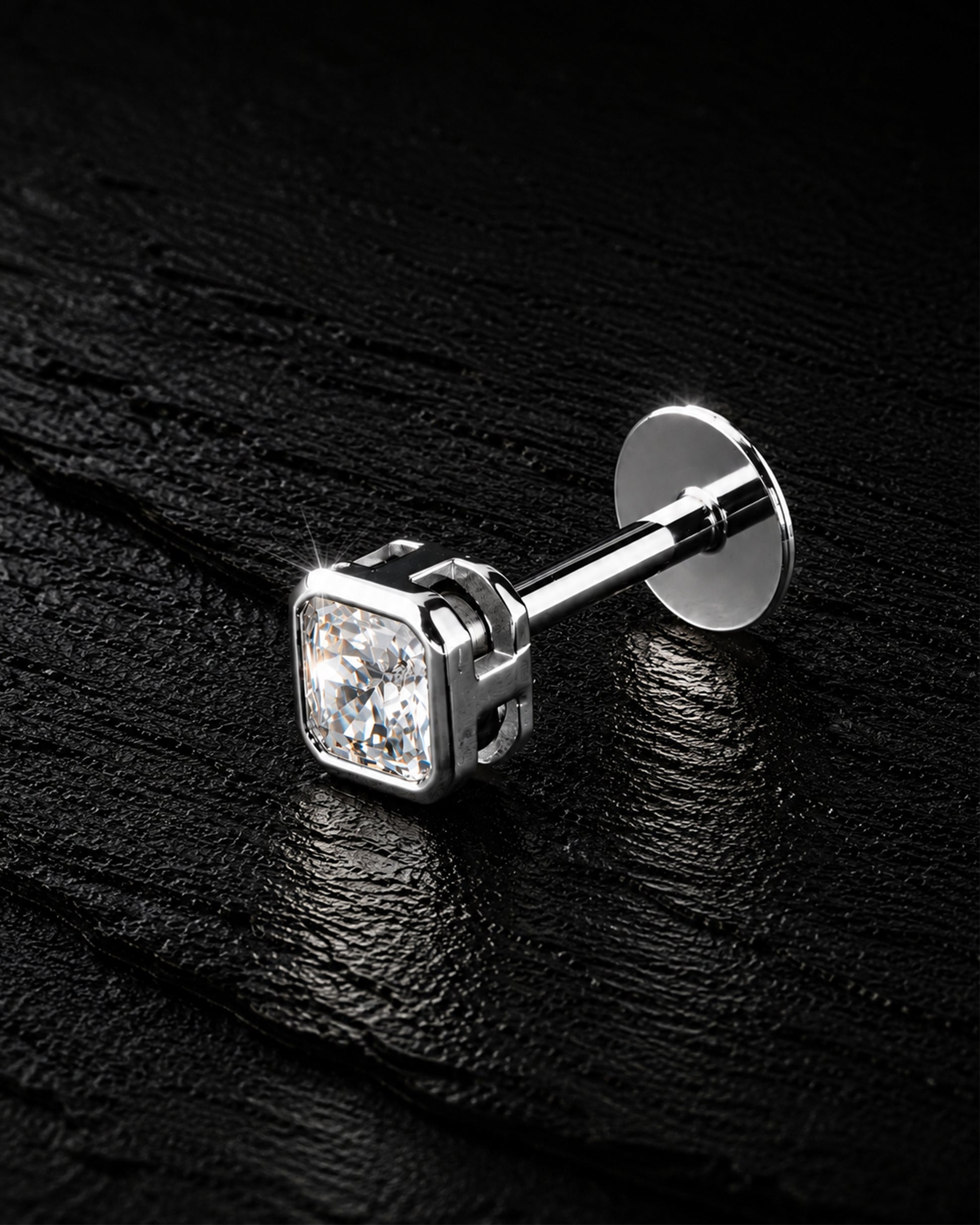 A single silver stud earring with a square-cut clear gemstone, displayed on a textured black background.