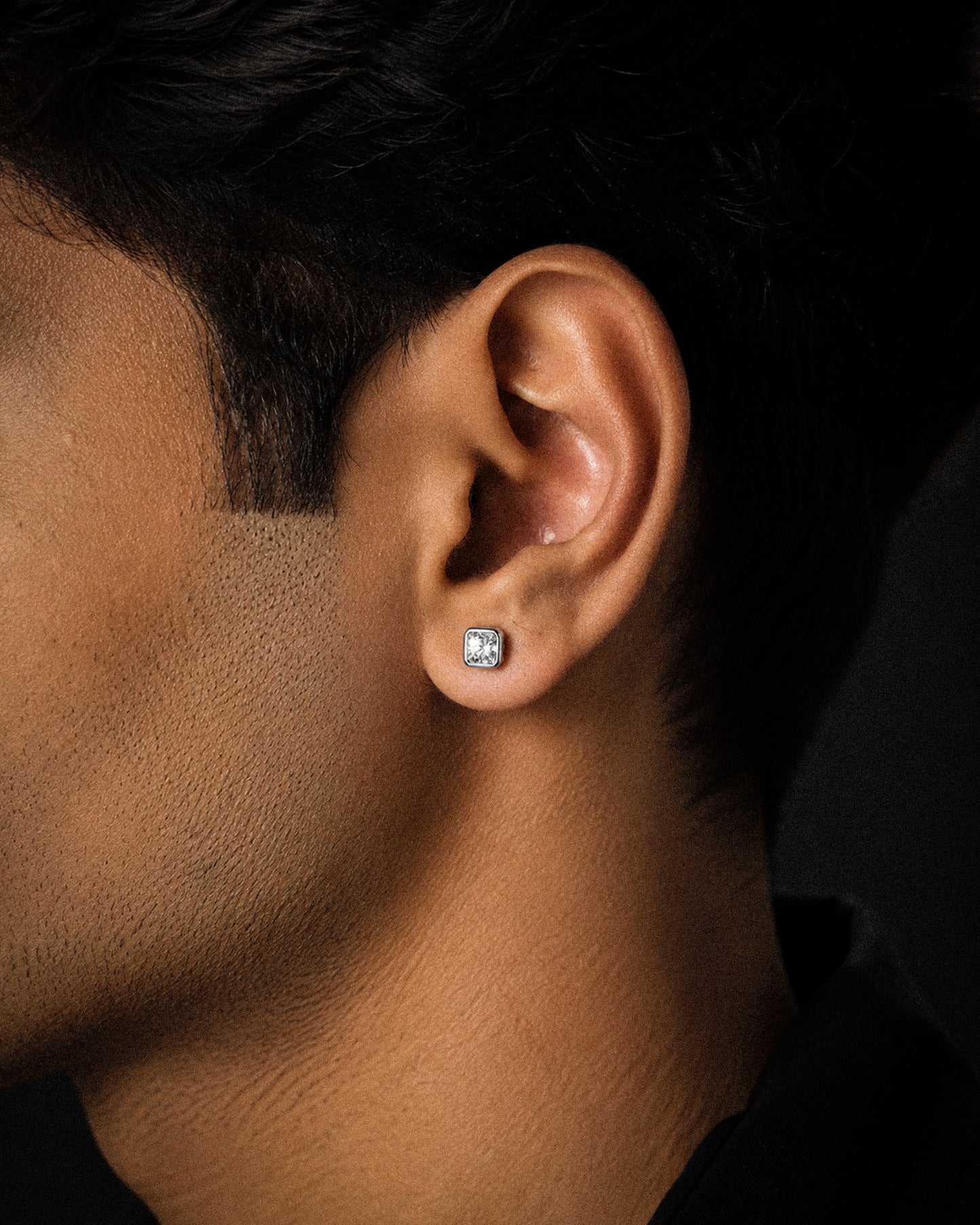 A close-up side profile of a man's ear wearing a small square stud earring with a clear gemstone in a silver-toned setting, against a dark background.