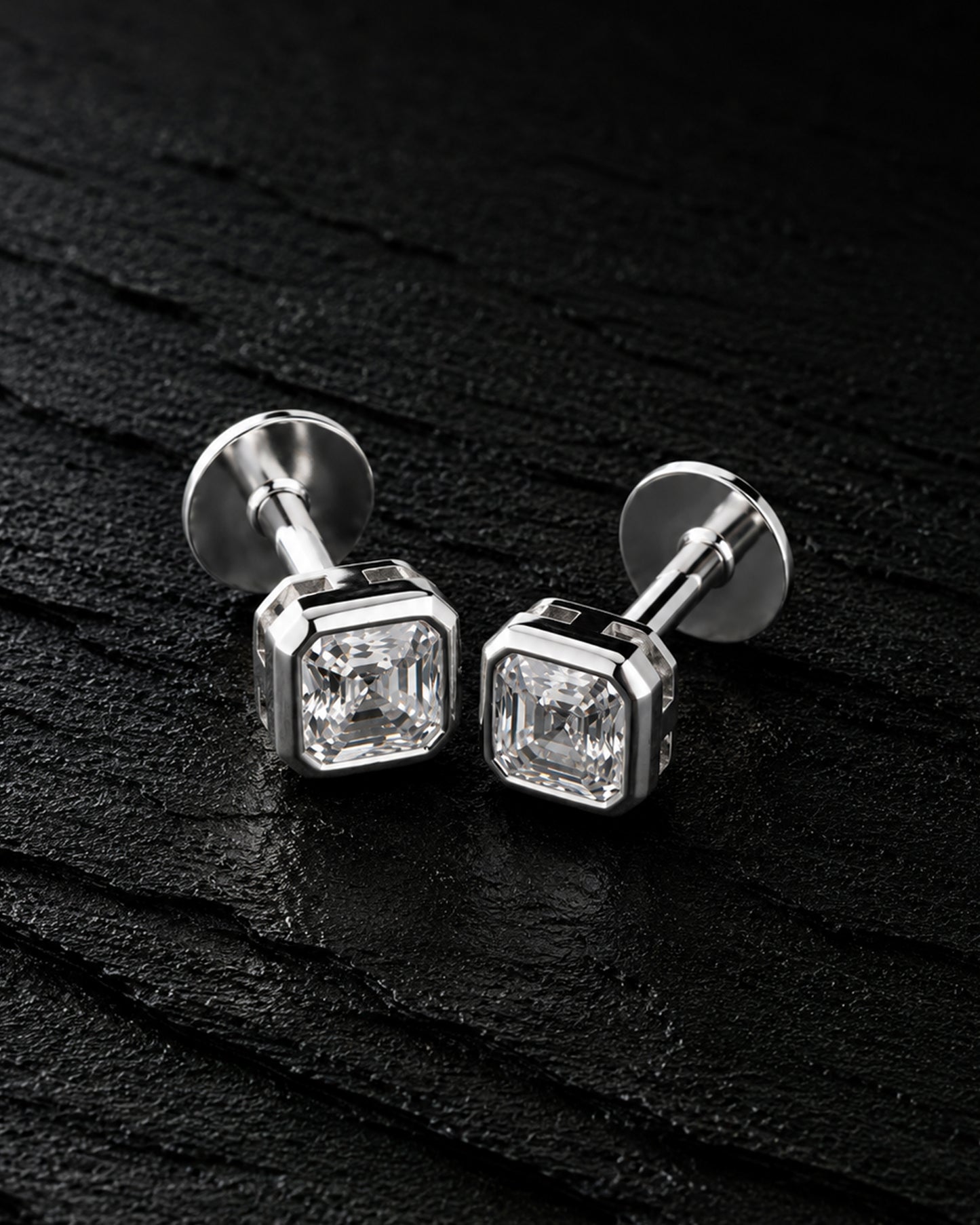 A pair of silver-toned stud earrings with square bezel-set clear gemstones, displayed on a dark textured surface.