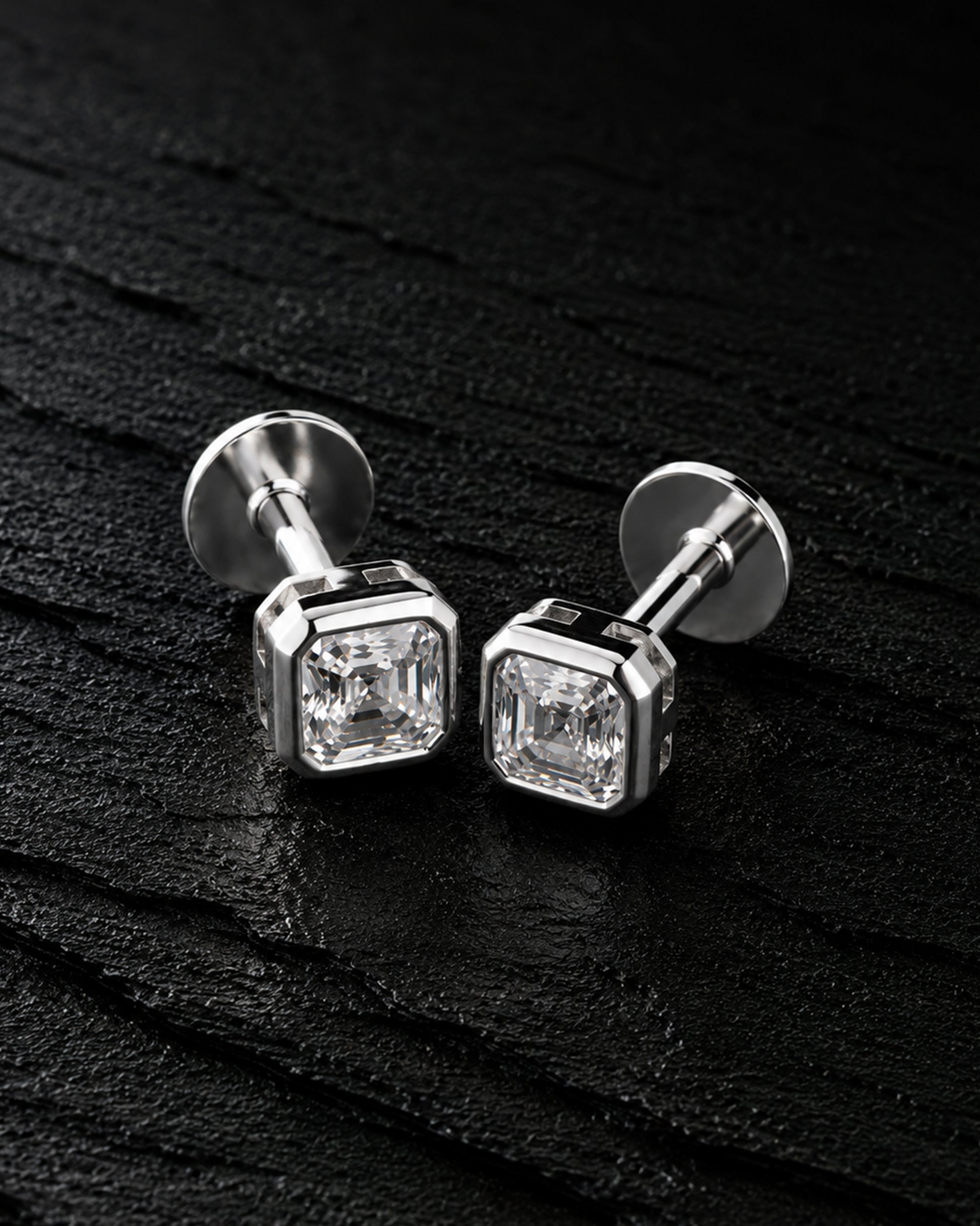 A pair of silver-toned stud earrings with square bezel-set clear gemstones, displayed on a dark textured surface.