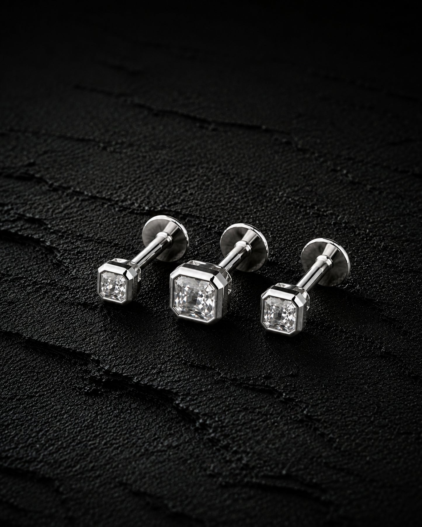 Three silver stud earrings with square-cut clear gemstones, arranged on a dark textured surface.