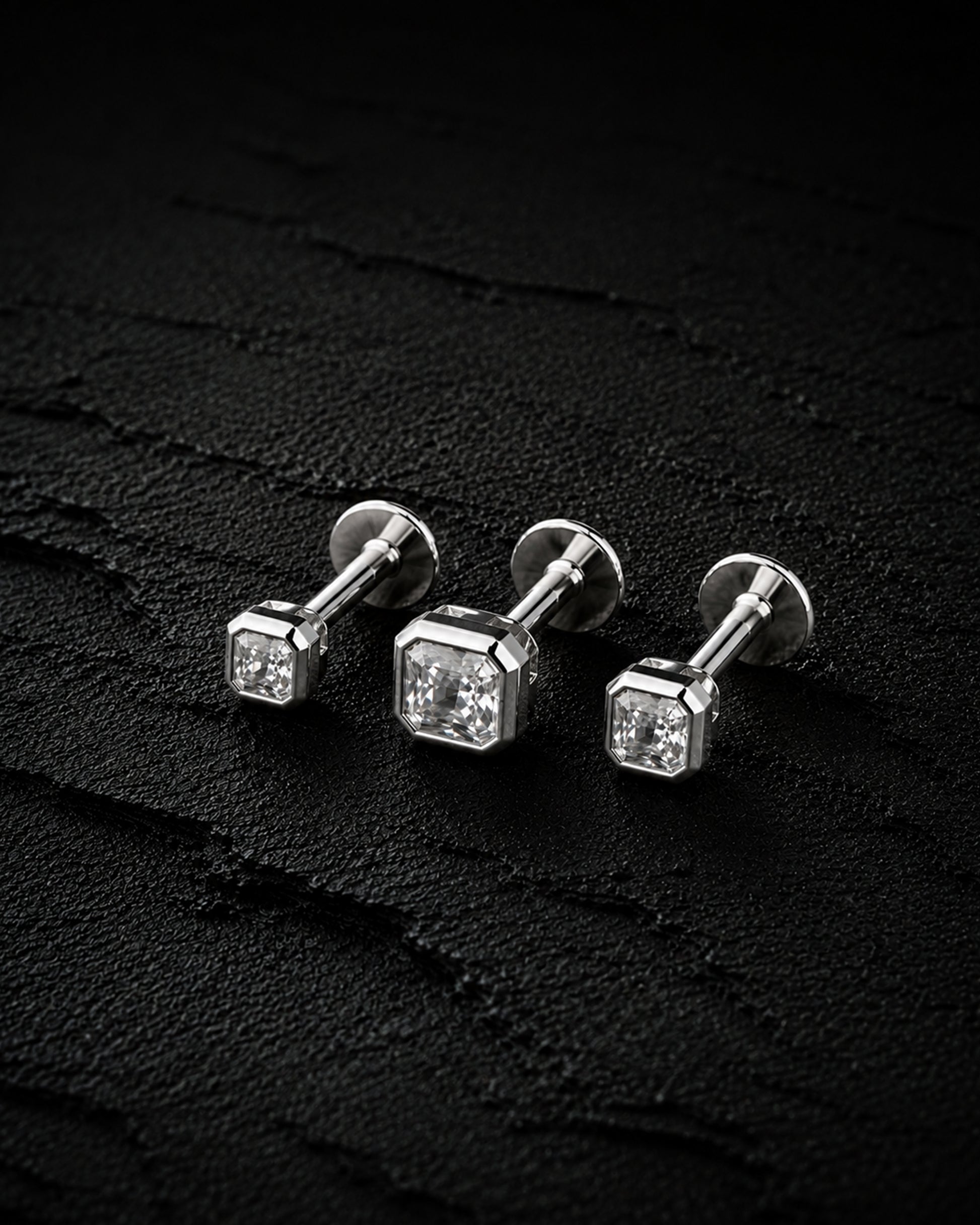 Three silver stud earrings with square-cut clear gemstones, arranged on a dark textured surface.