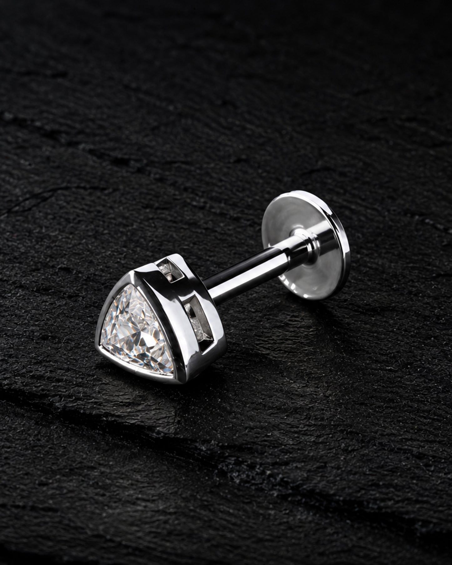 A silver-toned triangular stud earring with a clear faceted gemstone, shown on a dark textured background.