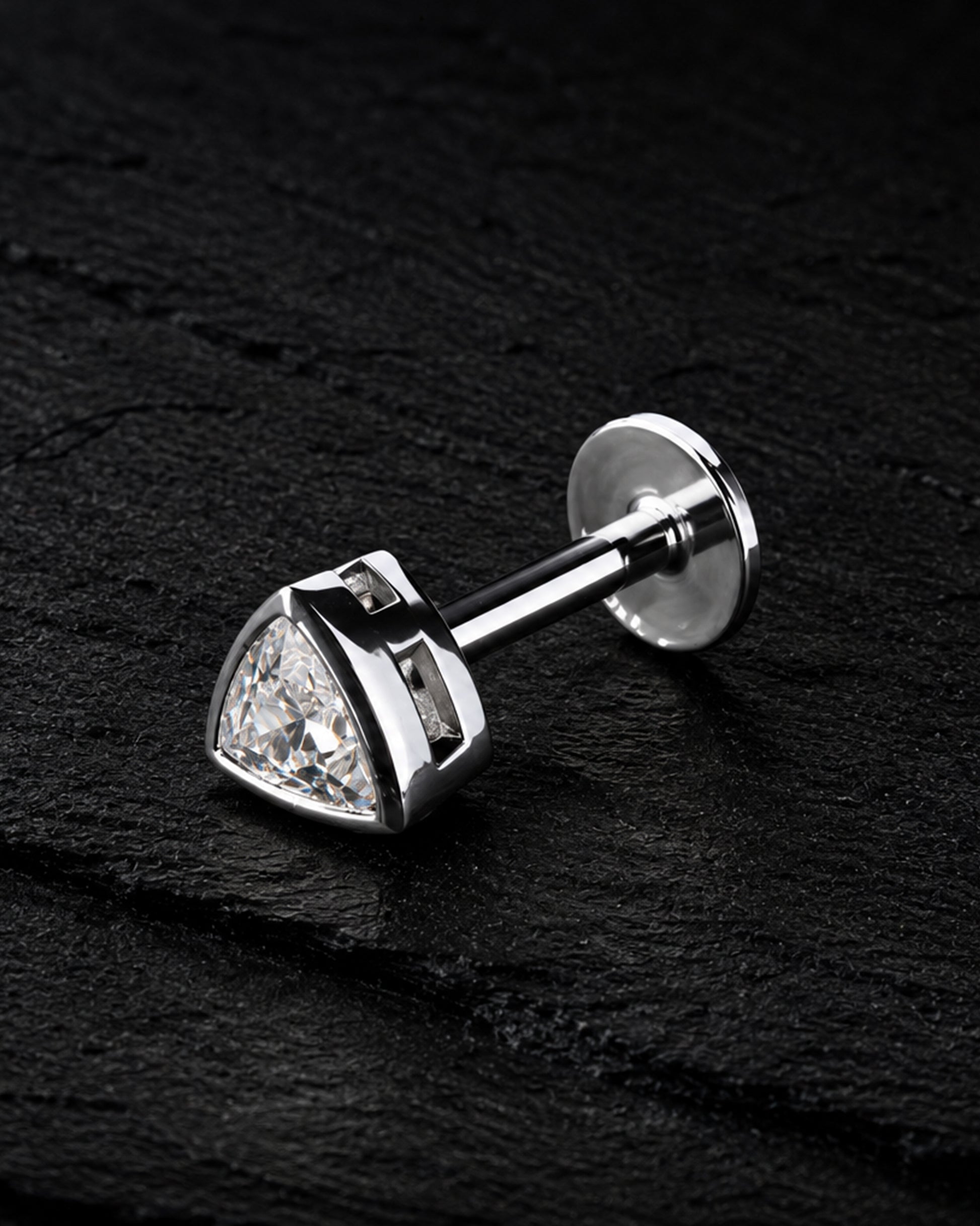 A silver-toned triangular stud earring with a clear faceted gemstone, shown on a dark textured background.