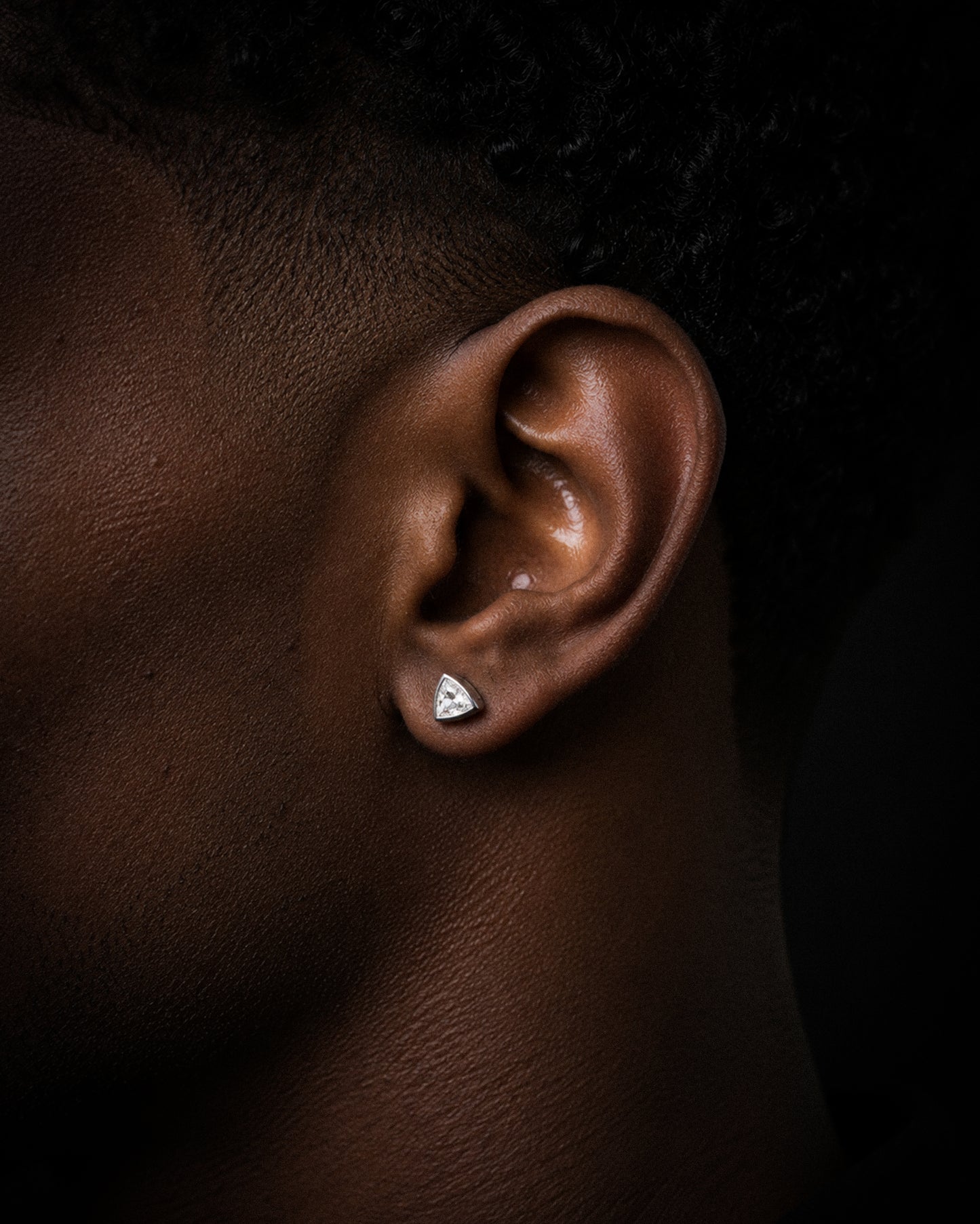 A close-up side profile of a person's ear wearing a small triangular silver stud earring against a dark background.