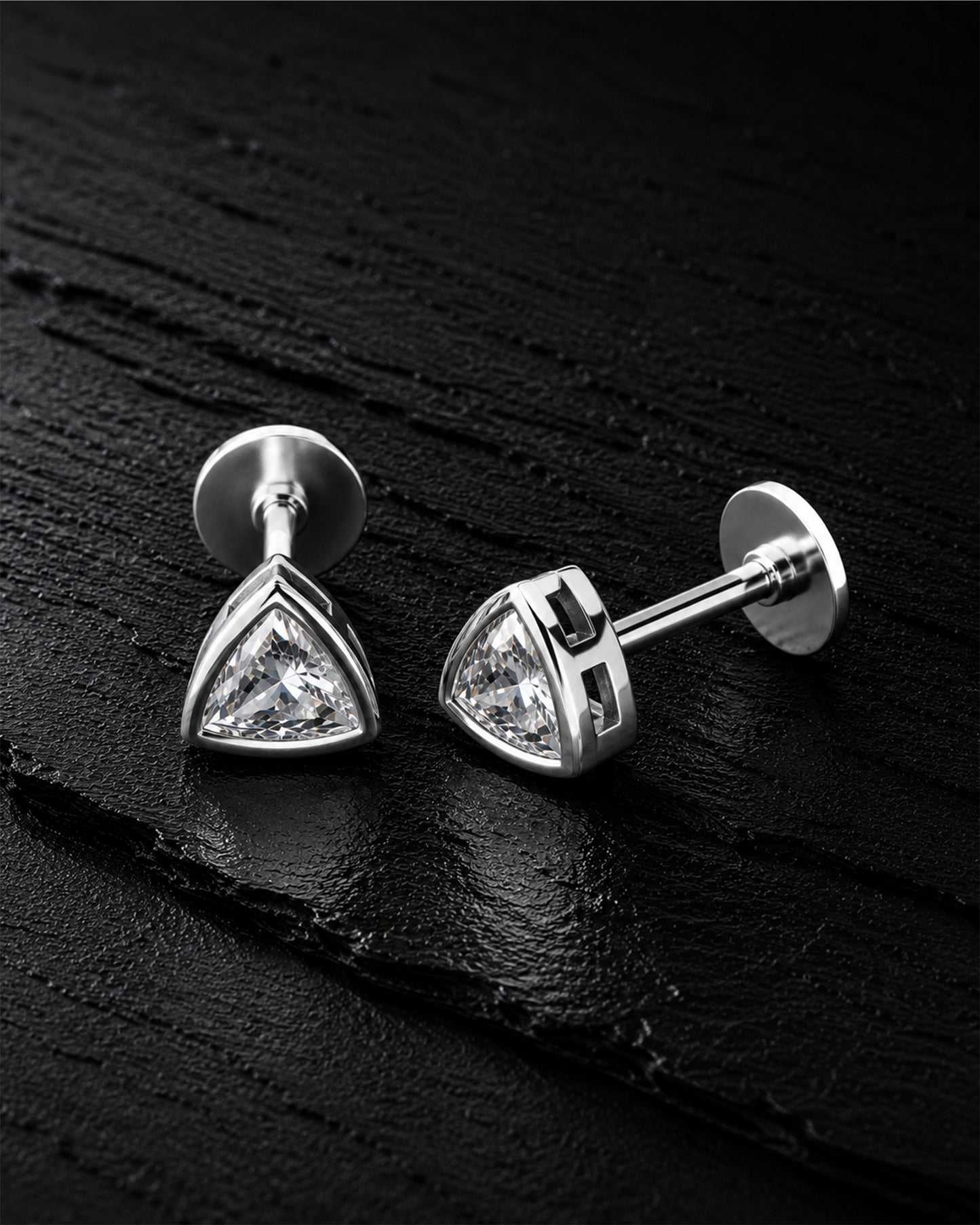 A pair of silver triangular stud earrings with clear gemstones, displayed on a dark textured background.