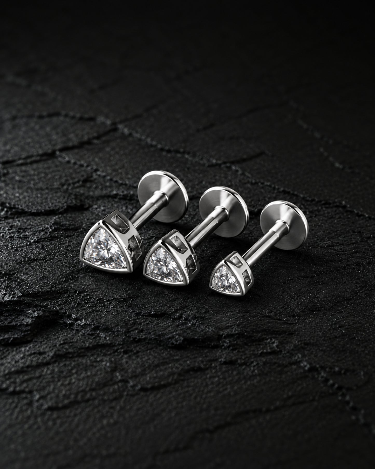 Three silver-toned triangular stud earrings with clear stones, arranged on a dark textured surface.