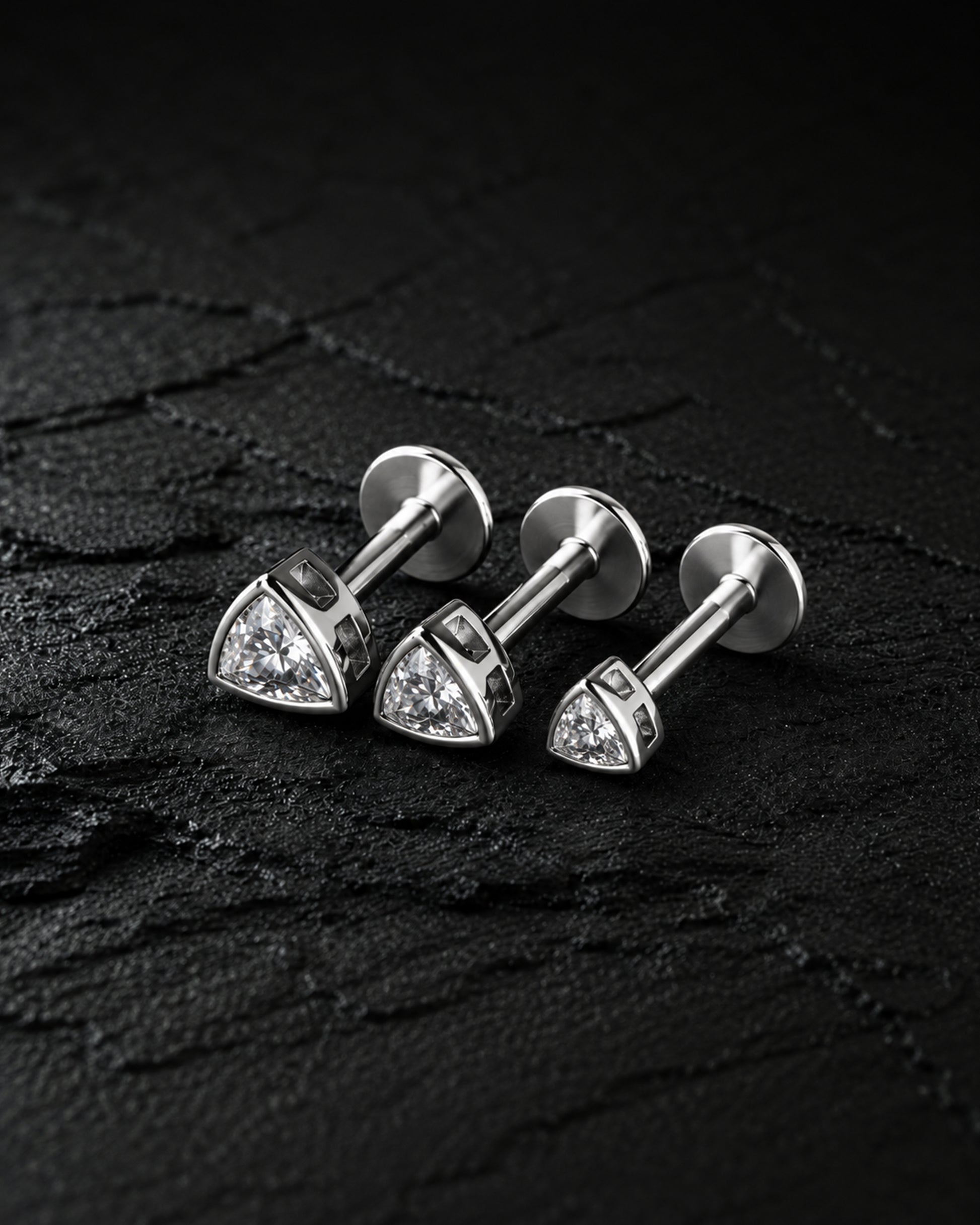 Three silver-toned triangular stud earrings with clear stones, arranged on a dark textured surface.