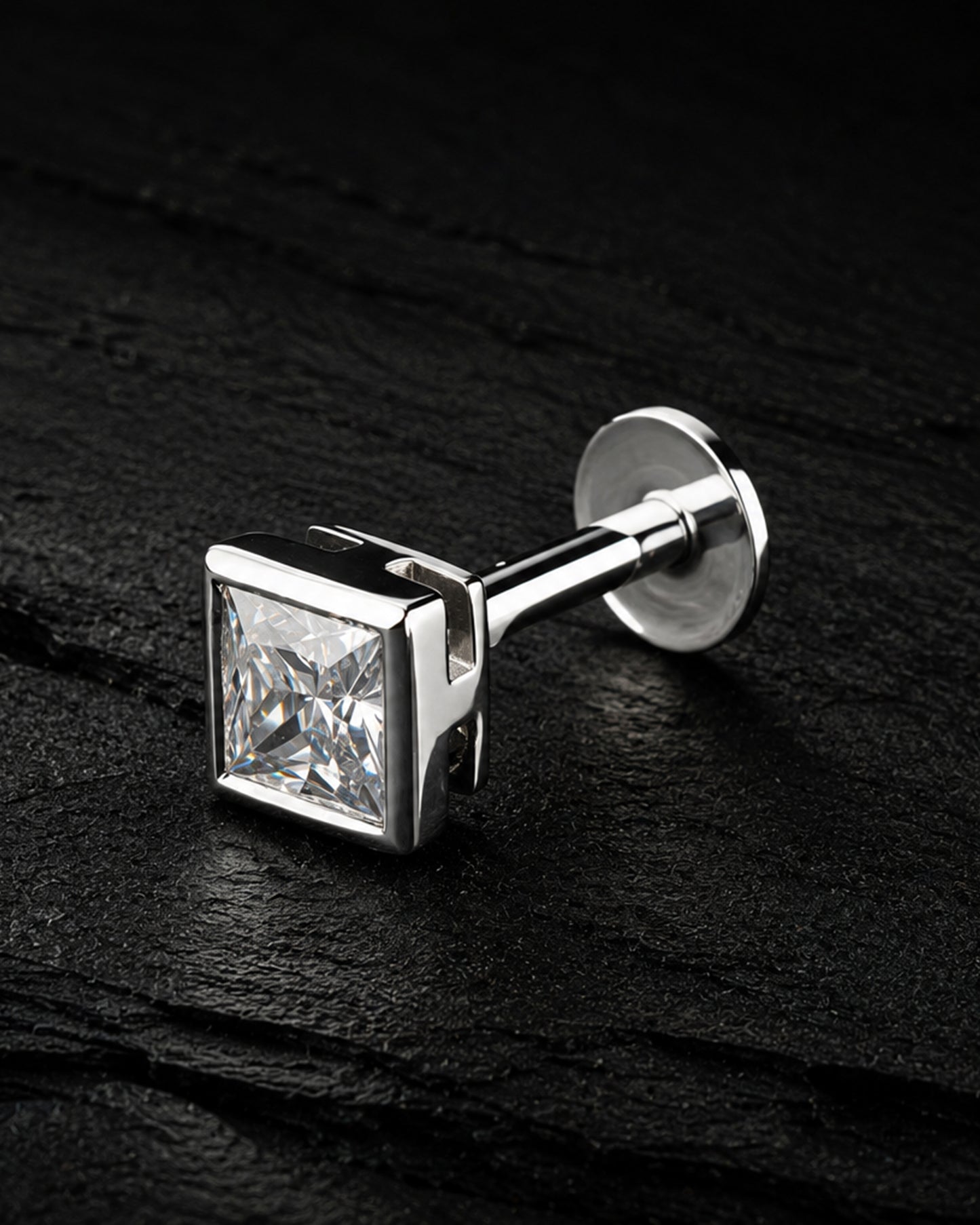 A silver-toned men's cufflink featuring a square princess-cut clear gemstone, displayed on a dark textured slate surface.