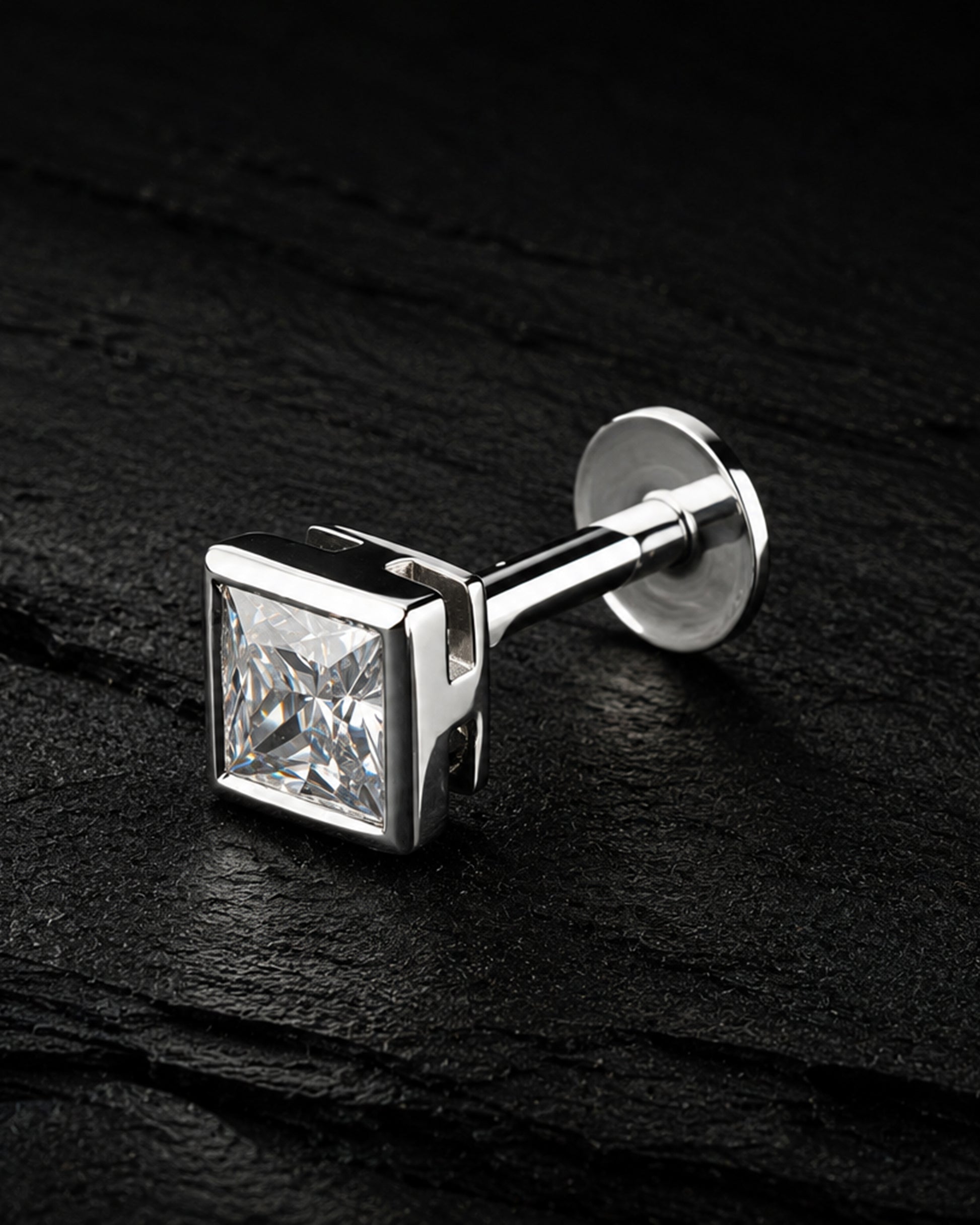 A silver-toned men's cufflink featuring a square princess-cut clear gemstone, displayed on a dark textured slate surface.