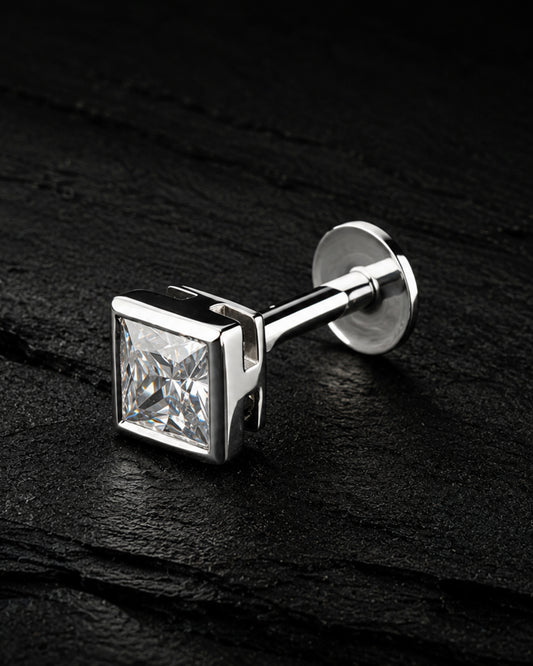 A silver-toned men's cufflink featuring a square princess-cut clear gemstone, displayed on a dark textured slate surface.