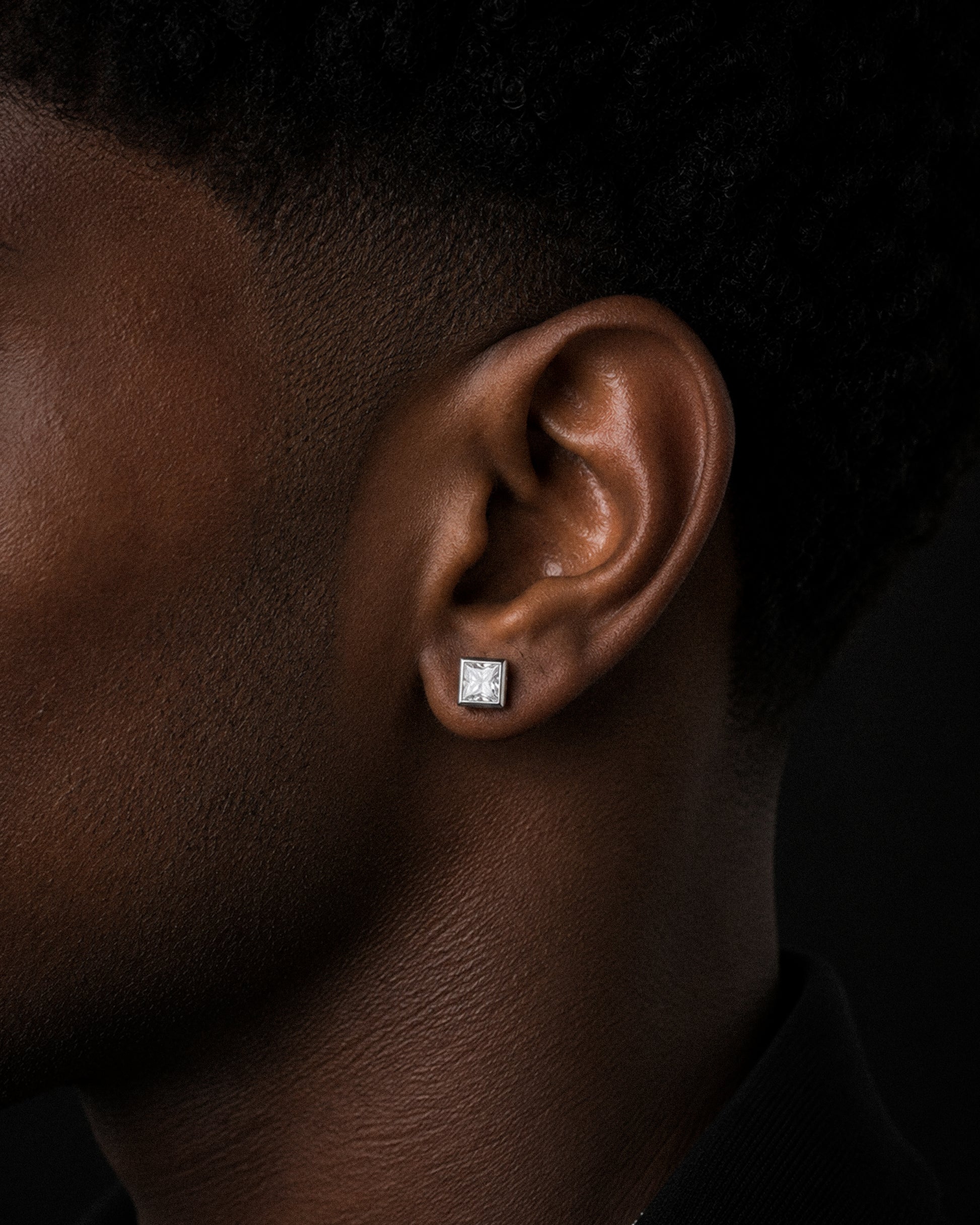 A close-up side profile of a person's ear wearing a small square stud earring with a clear gemstone in a silver-toned setting, against a dark background.