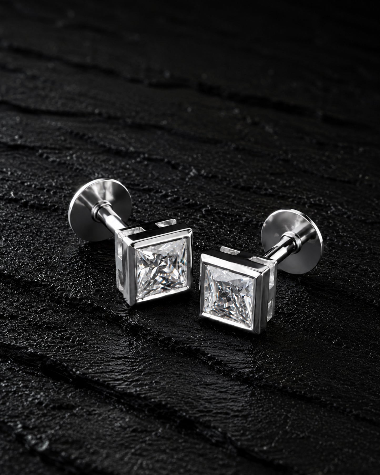 A pair of silver-toned square stud earrings with clear princess-cut gemstones, displayed on a dark textured surface.