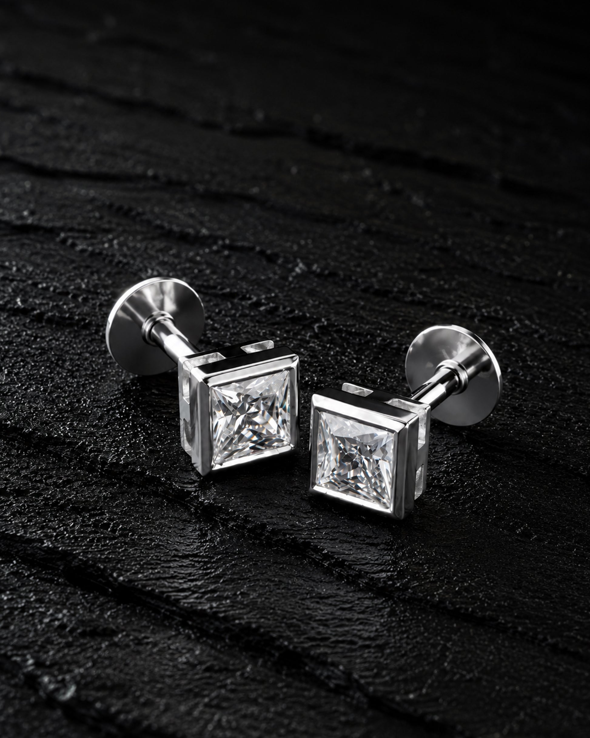 A pair of silver-toned square stud earrings with clear princess-cut gemstones, displayed on a dark textured surface.
