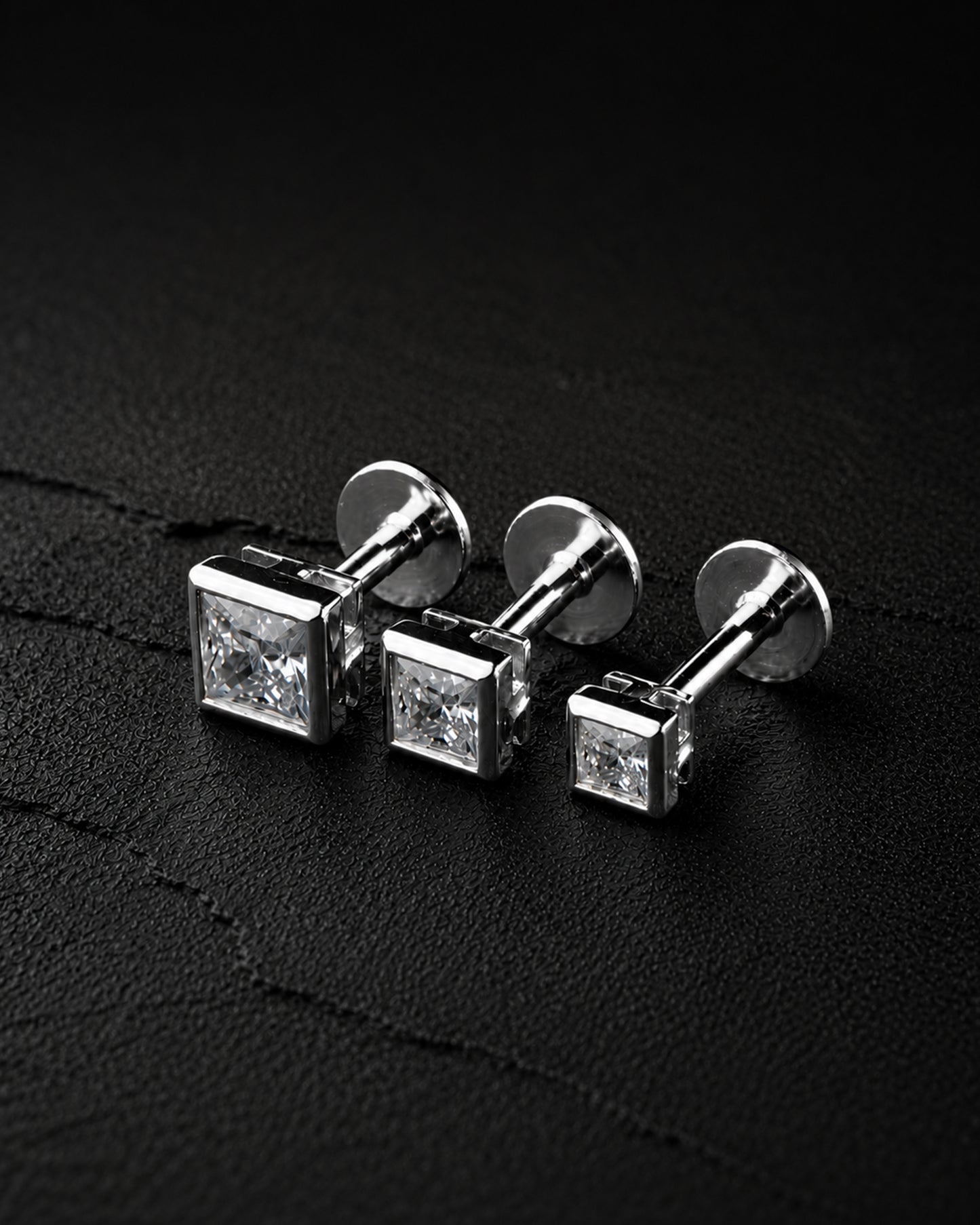 Three silver-toned square stud earrings with clear gemstones, arranged on a dark textured surface.