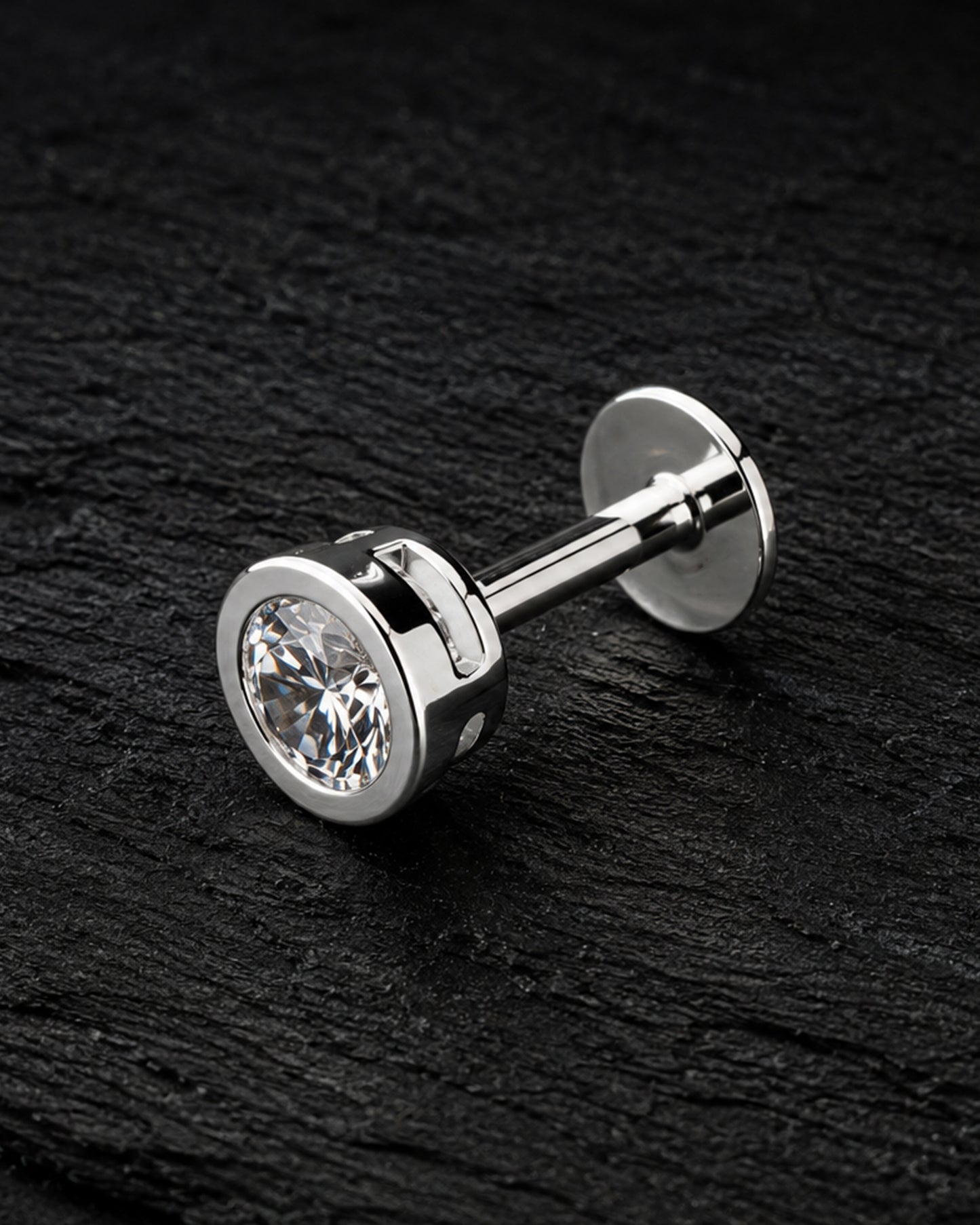 A pair of silver-toned labret studs with round bezel-set clear gemstones, displayed diagonally on a dark textured slate surface.