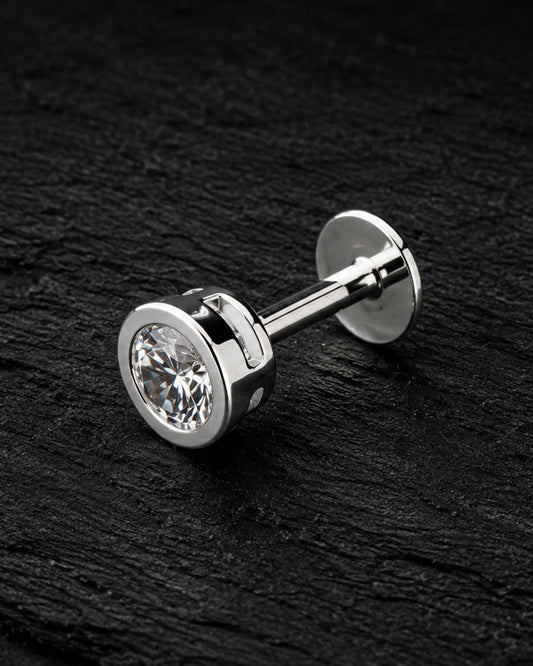 A pair of silver-toned labret studs with round bezel-set clear gemstones, displayed diagonally on a dark textured slate surface.