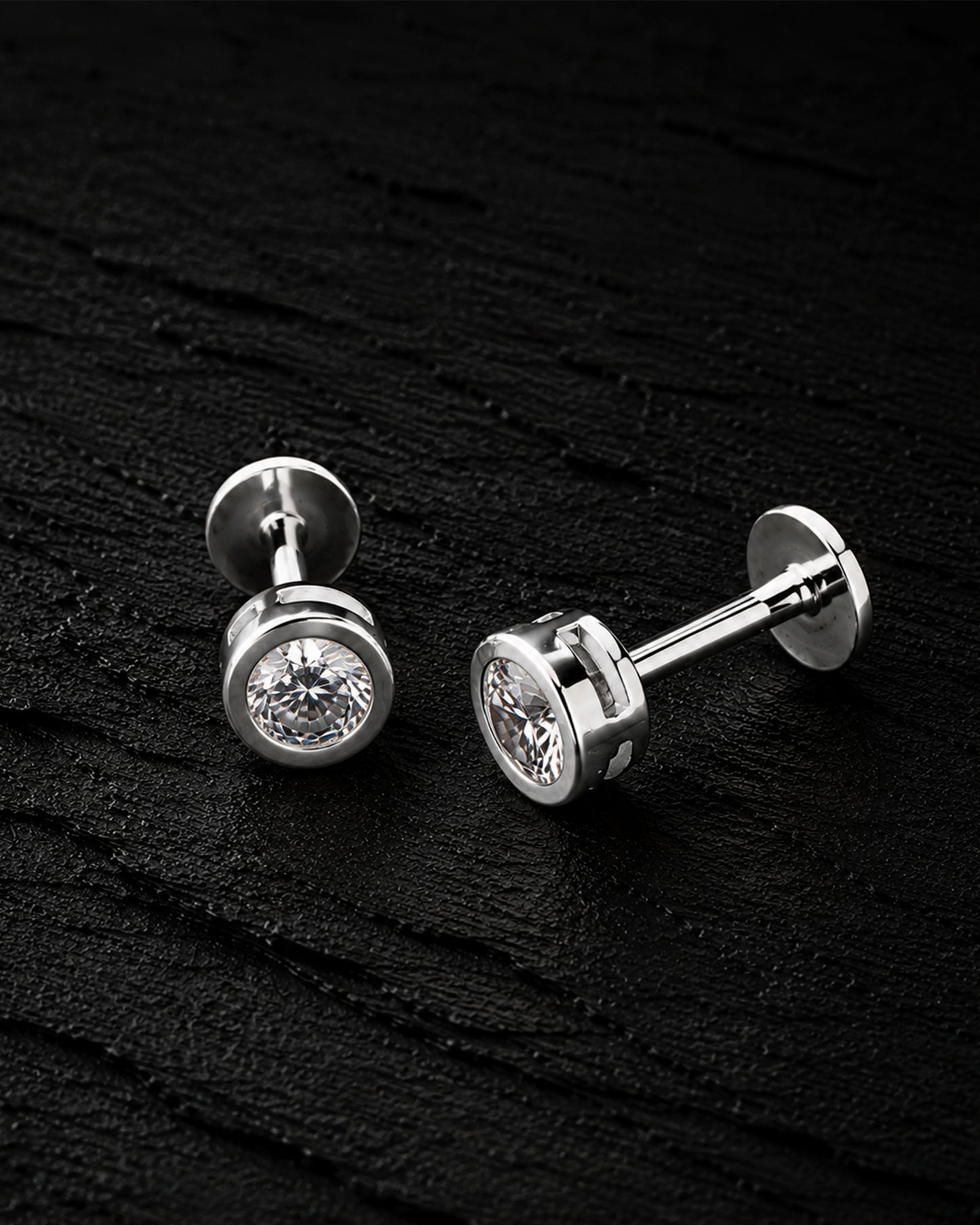 A pair of silver-toned stud earrings with round bezel-set clear gemstones, displayed on a dark textured background.