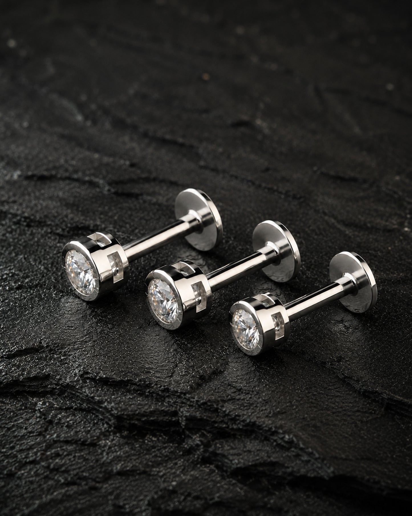 Three silver-toned labret stud earrings with round clear gemstones, arranged on a dark textured stone surface.
