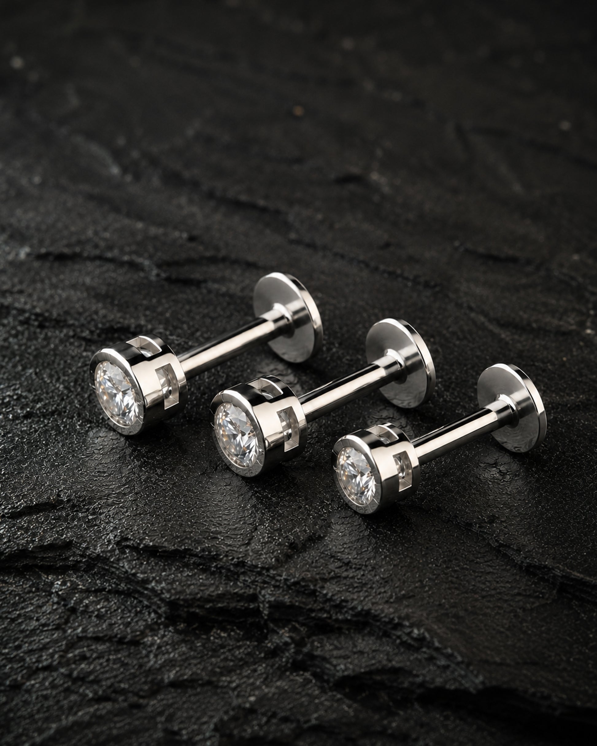Three silver-toned labret stud earrings with round clear gemstones, arranged on a dark textured stone surface.